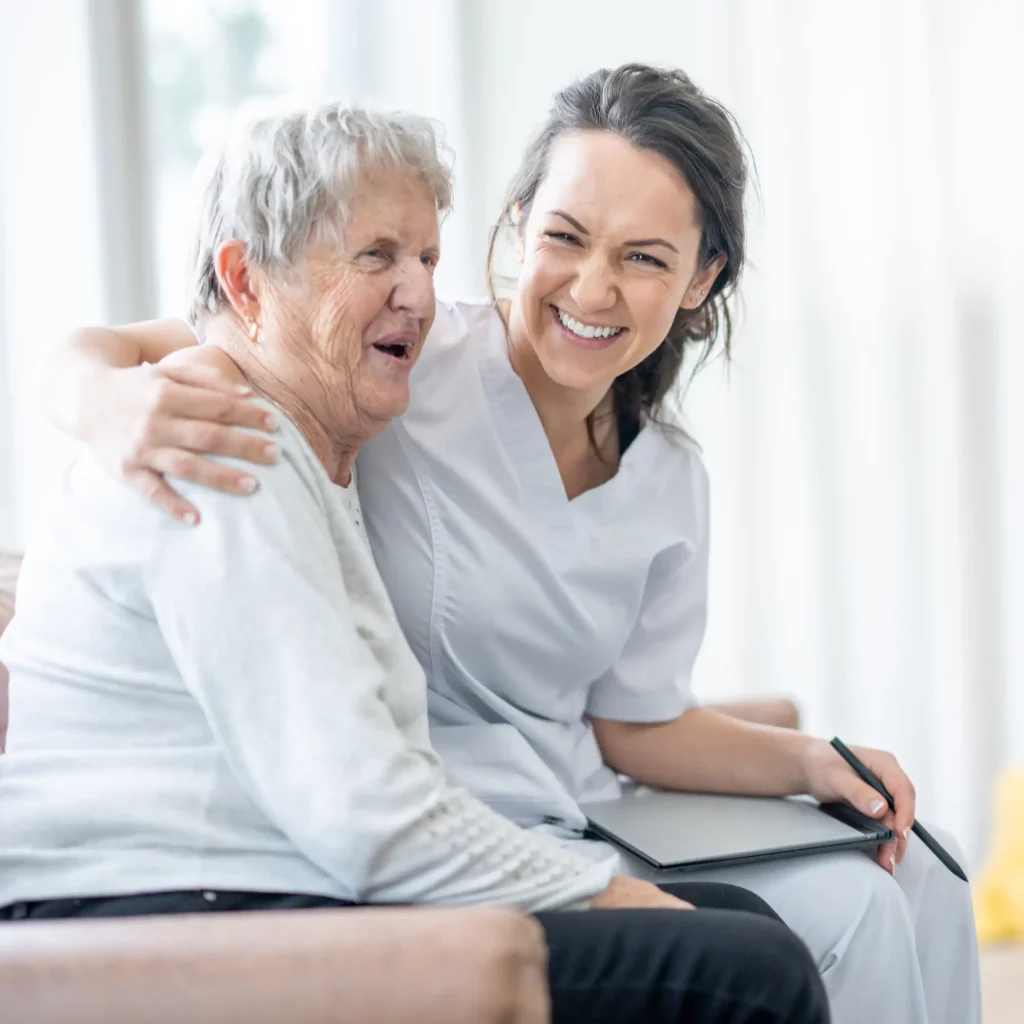 Pflegekraft und Seniorin sitzen lachend nebeneinander
