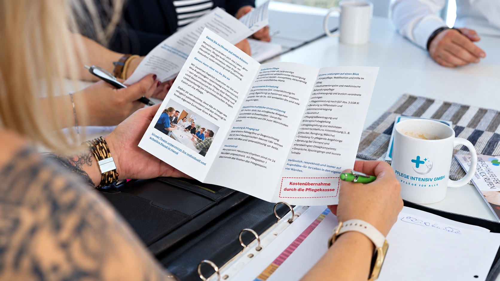 Hände halten einen aufgeklappten Flyer zu Pflegeleistungen am Besprechungstisch